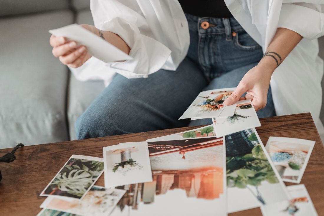 A person's hands holding a small printed photograph above a wooden table scattered with other prints of plants and buildings, face out of frame.