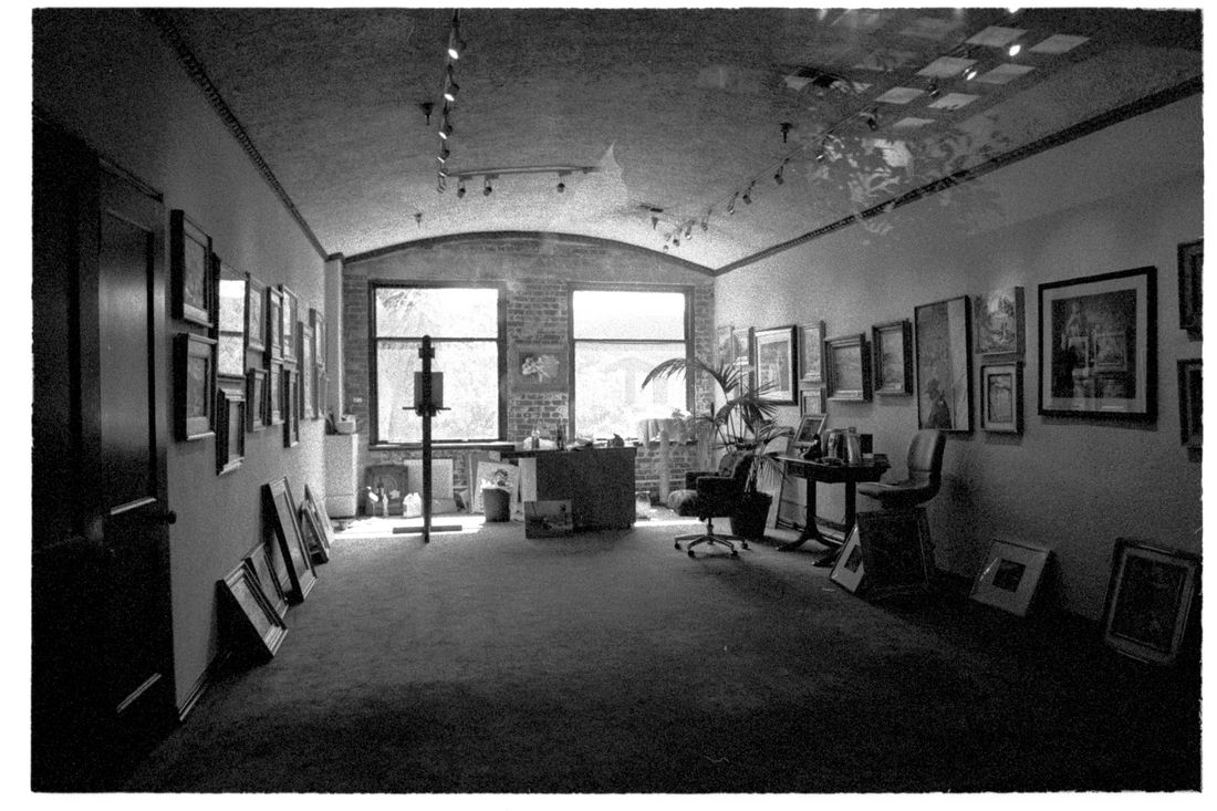 A black-and-white view of a working gallery room with framed pictures hung close together on both walls and more framed pieces leaning on the floor
