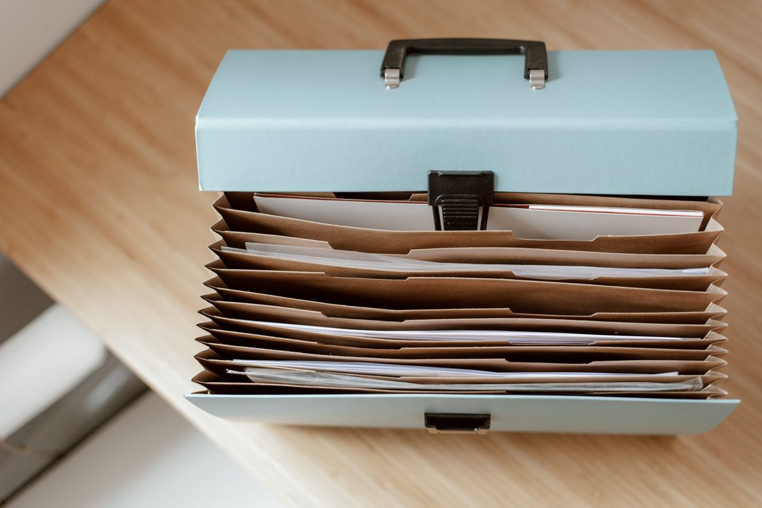 An open accordion file box of paper folders sitting on a light wooden desk