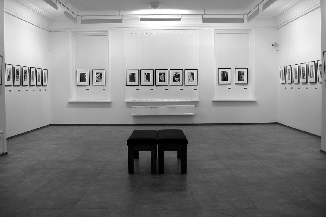 An empty gallery room with framed artwork on white walls and two low benches on the floor