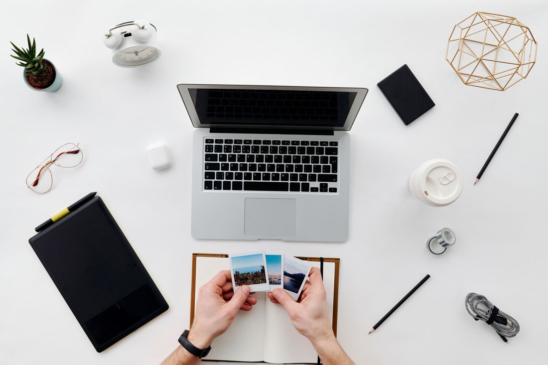 Overhead view of hands holding small printed photographs next to an open laptop on a plain desk