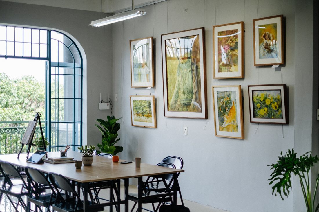 A room with a long table and a wall of framed paintings of varying sizes lit by natural window light