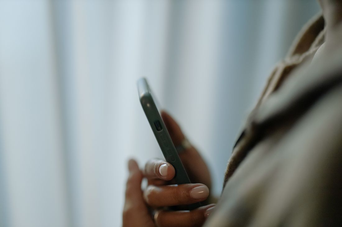 A hand holding a phone in profile with the screen turned to the side and unreadable, against a softly blurred plain curtain background.