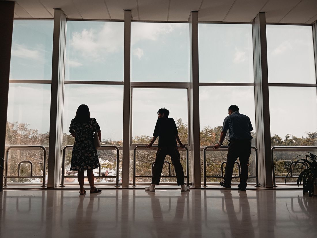 Three people seen from behind, standing apart along a tall window in a bright interior