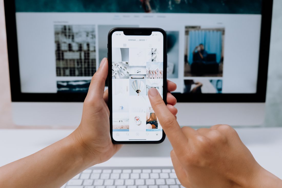 Hands holding a smartphone open to a social media app in front of a desktop computer