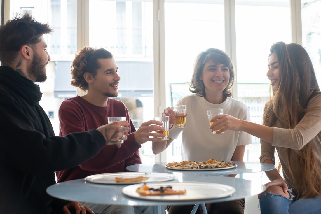 Four friends laughing and toasting drinks around a small table with pizza in a bright, casual indoor space