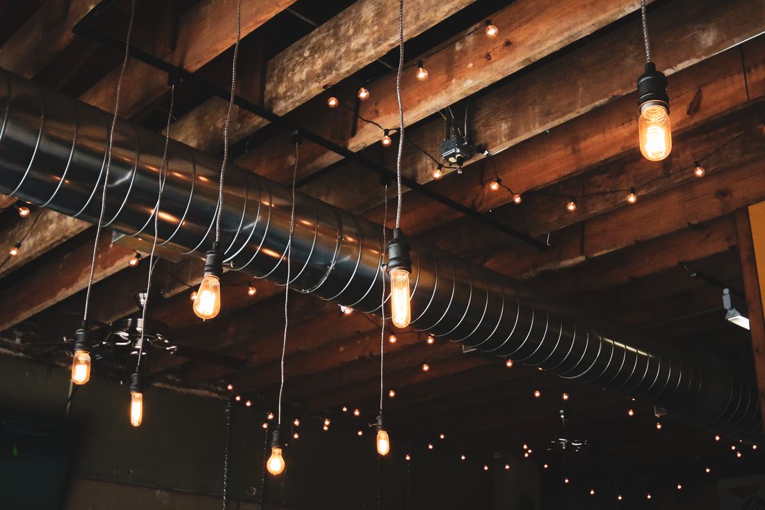 Warm string lights and hanging bulbs against an exposed brick and wood beam ceiling with ductwork, nobody present