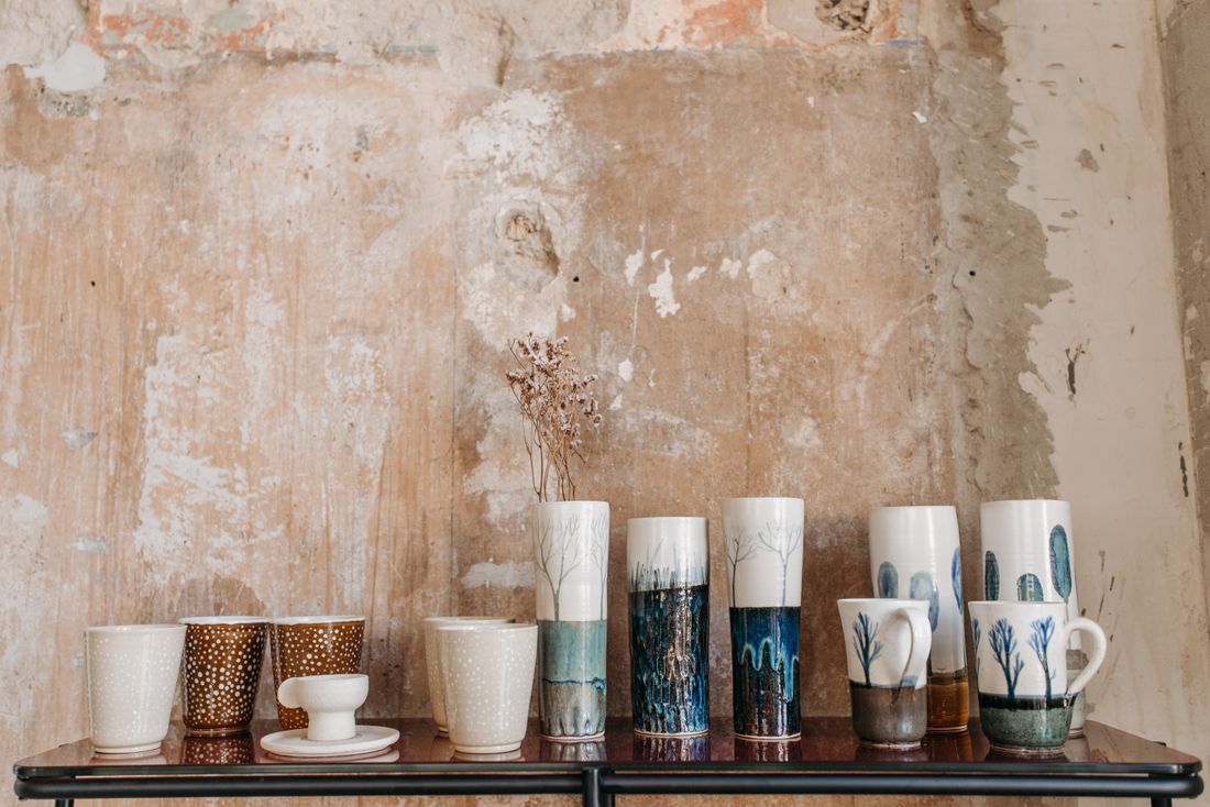 Handmade ceramic mugs and vases arranged with visible spacing on a wooden shelf against a weathered plaster wall.