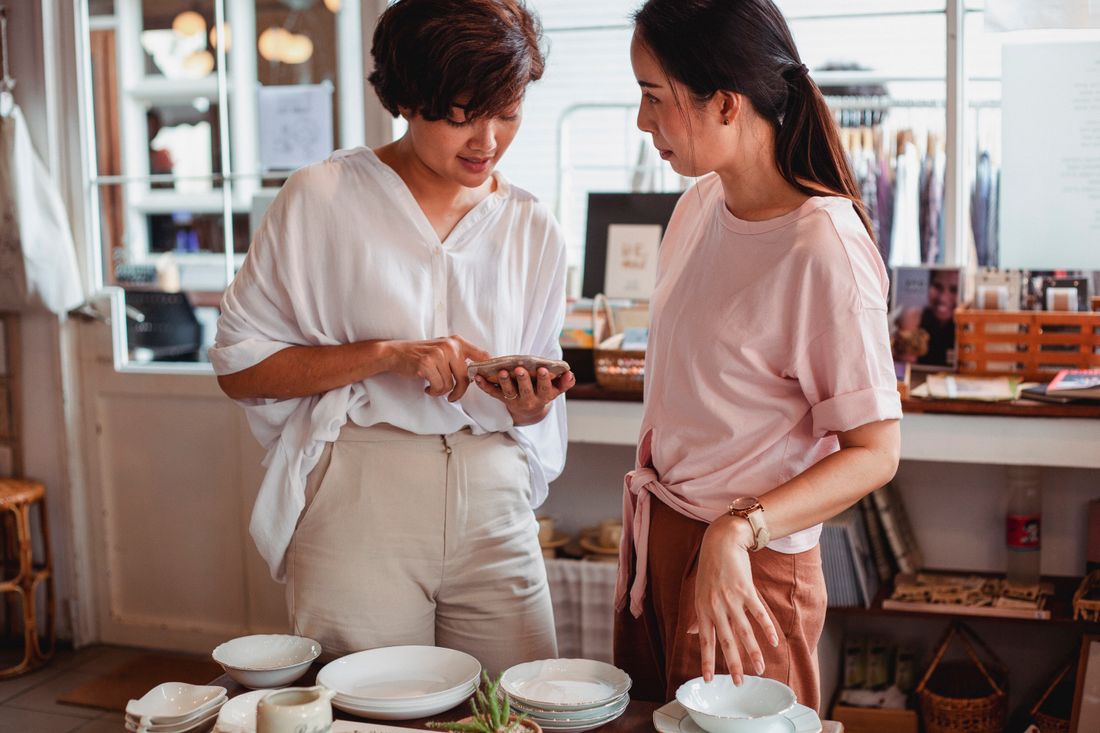 Two women standing at a shop counter looking closely at a small stack of ceramic dishware.