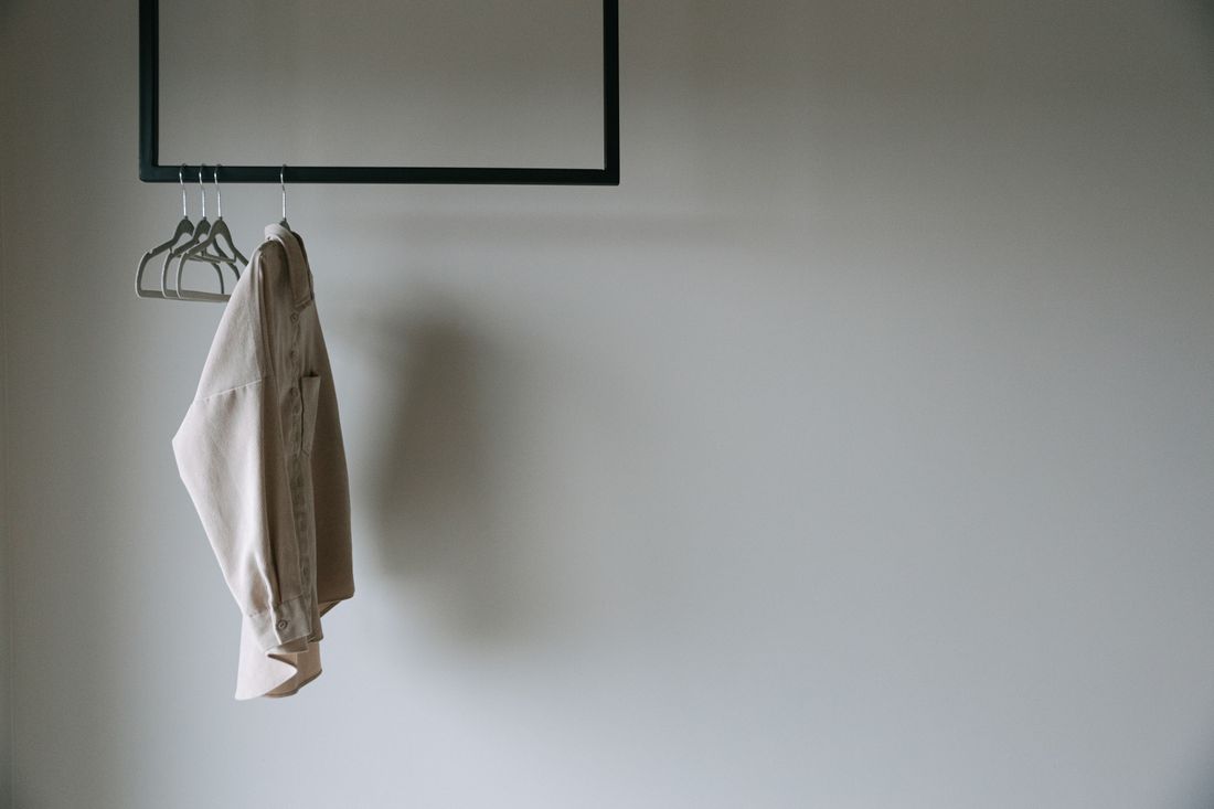 A single beige garment hanging on a minimal black wall-mounted rail beside a few empty hangers, against a plain wall in daylight.
