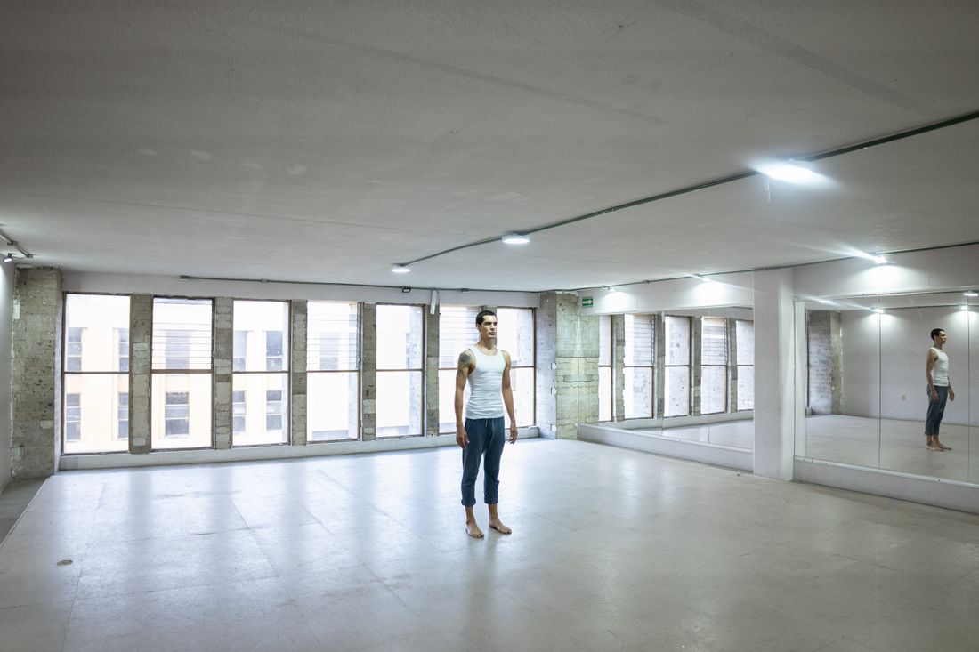 A person standing barefoot in an empty, bare-floored room with large windows, seen at a distance with no equipment in view.