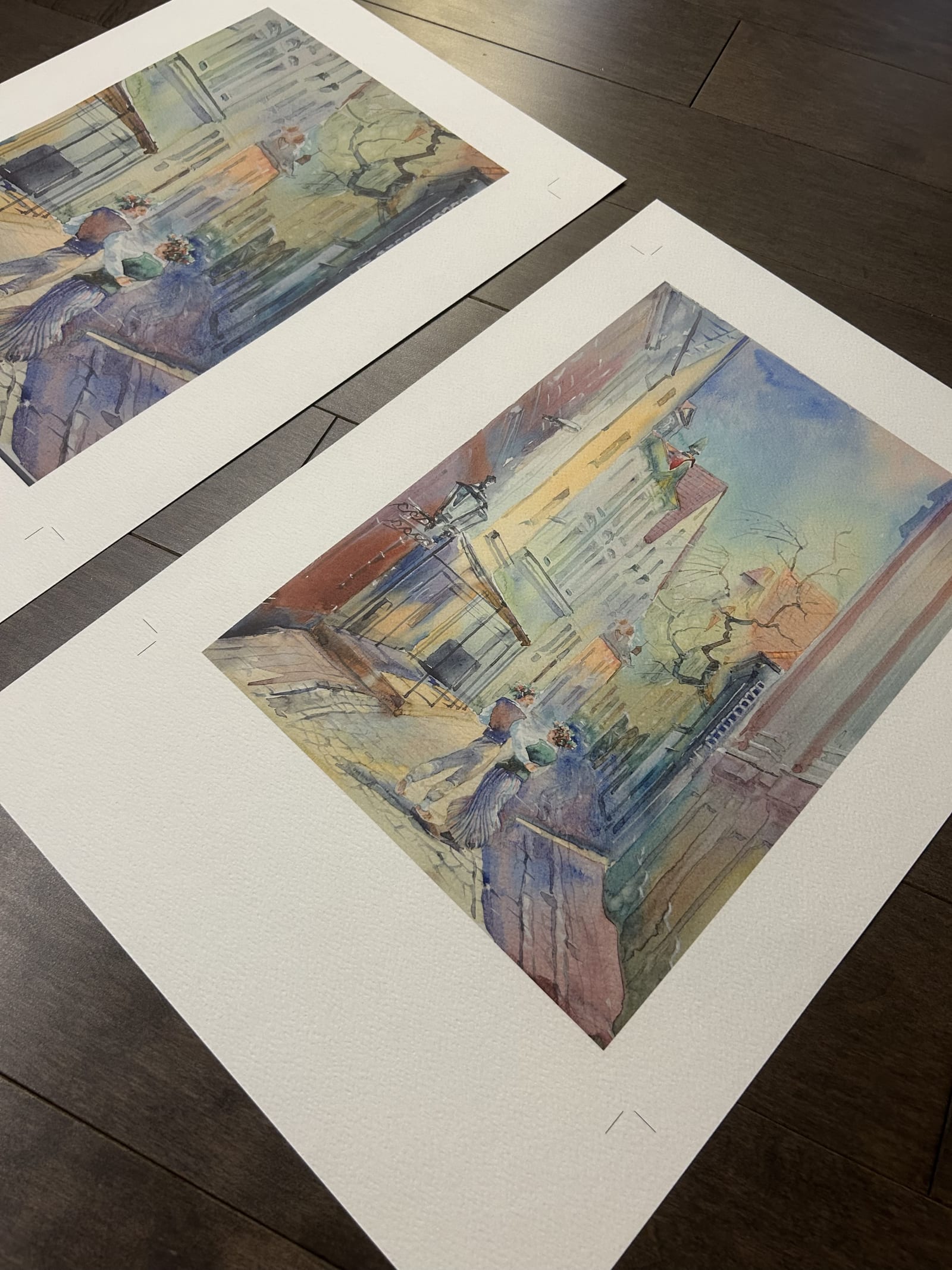 Two large watercolour prints of the same scene laid out on a studio floor for comparison during a proofing pass