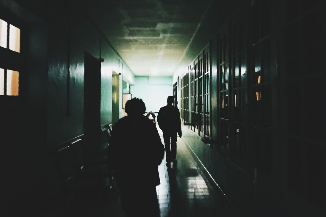 Two people seen from behind walking through a dim, plain institutional hallway.