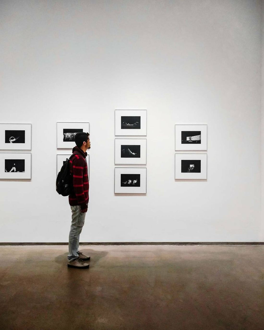 A person seen in profile standing back from a wall of framed photographs in a plain gallery space.