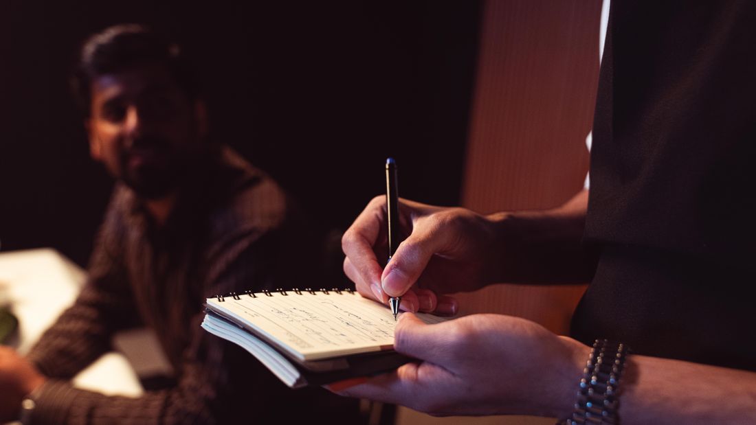 A close-up of hands writing notes in a small notebook during a conversation, with a second person blurred in the background.