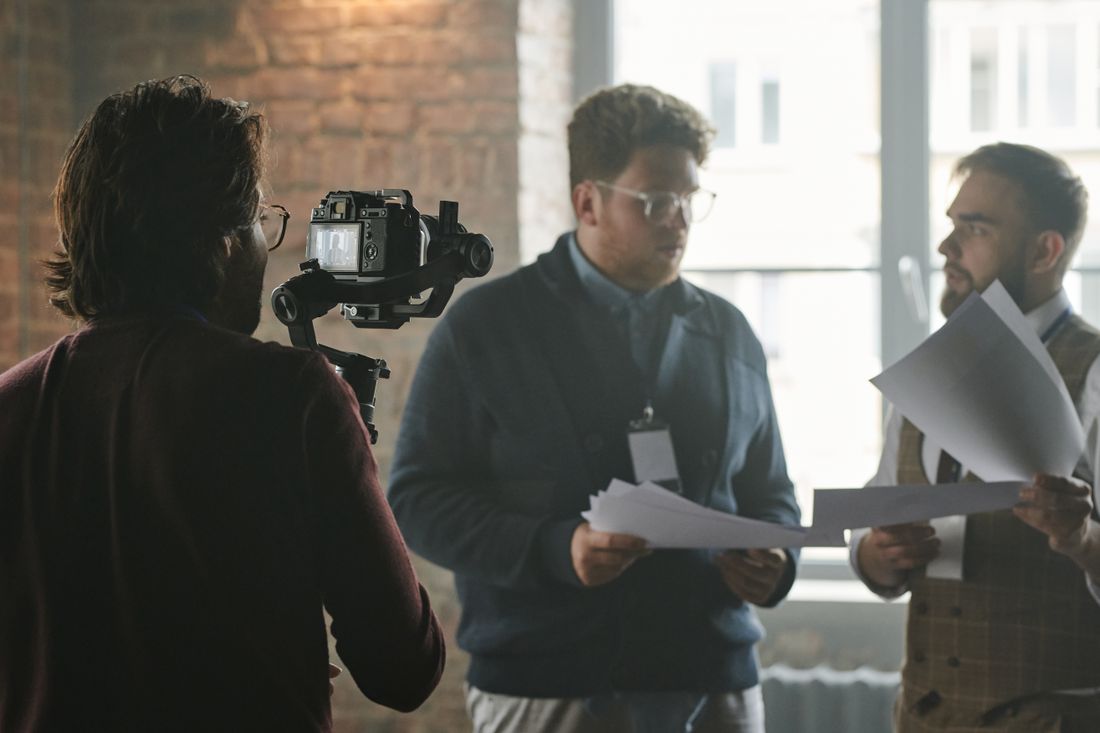A small crew filming two people in conversation indoors, with the camera operator visible from behind.