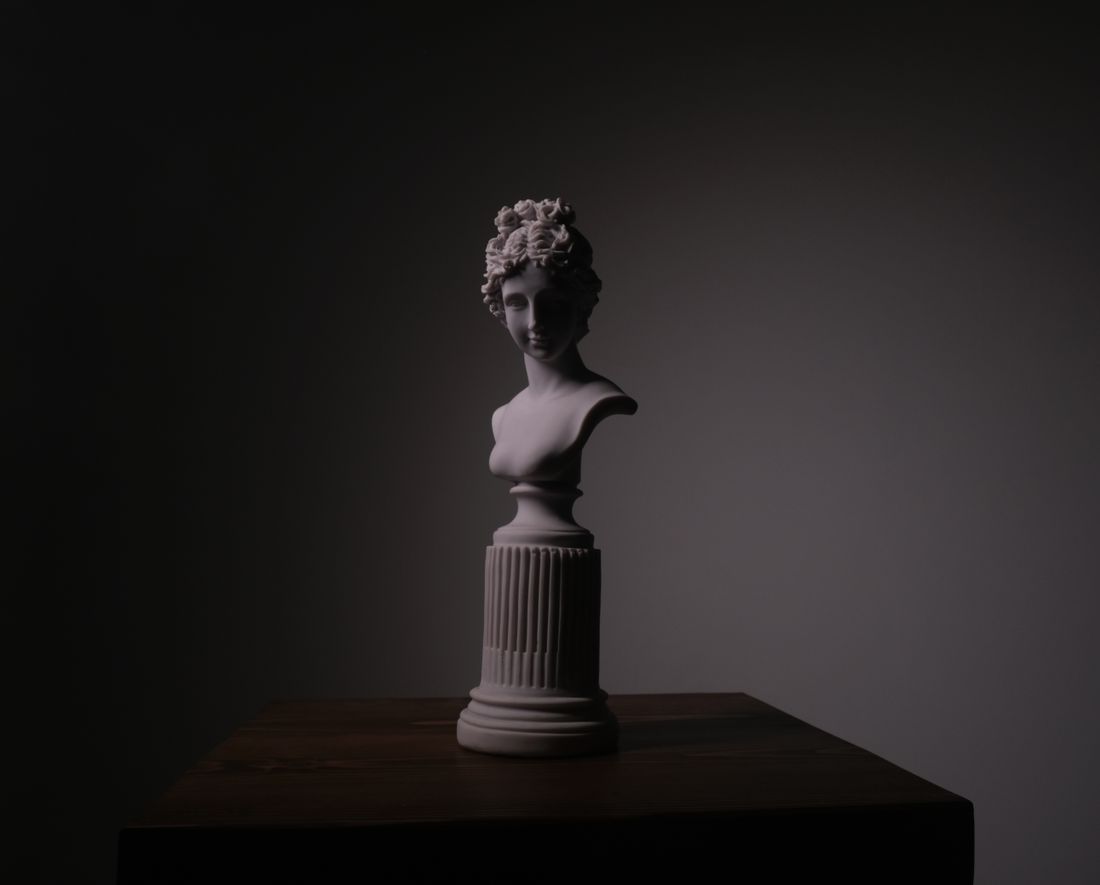 A plaster sculpture bust on a pedestal lit by a single directional studio light against a dark background