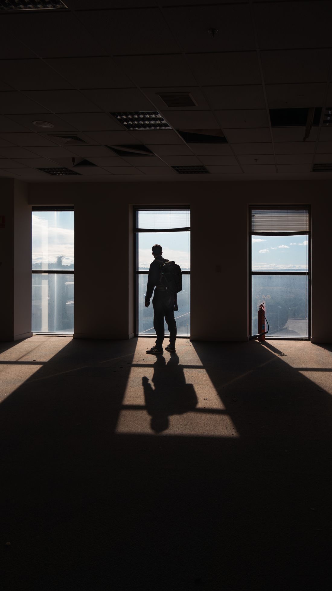 Silhouette of a person standing with their back to the camera at a tall office window, backpack over one shoulder.