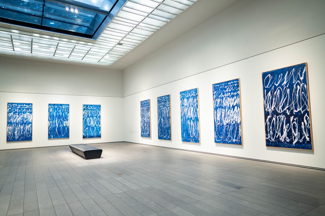 A gallery installation view of blue abstract paintings hung along white walls under a skylight