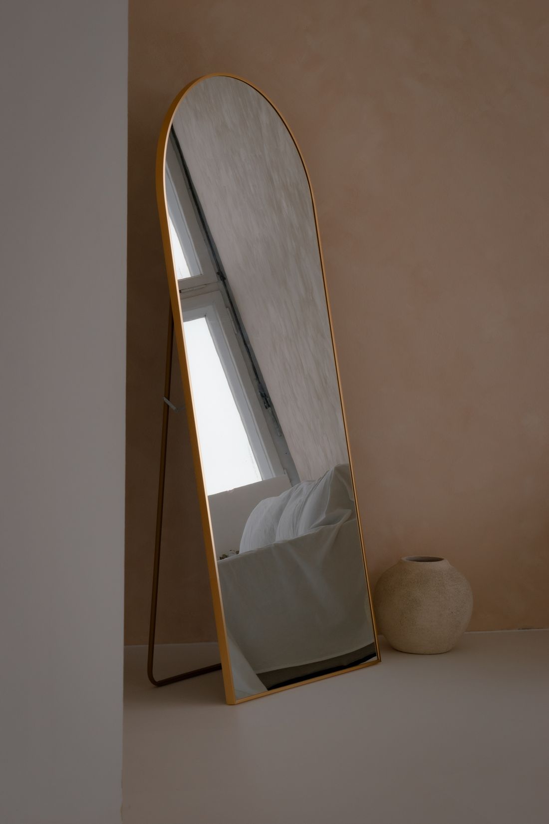 A standing arched mirror reflecting a softly lit bedroom corner with a window and a ceramic vase nearby