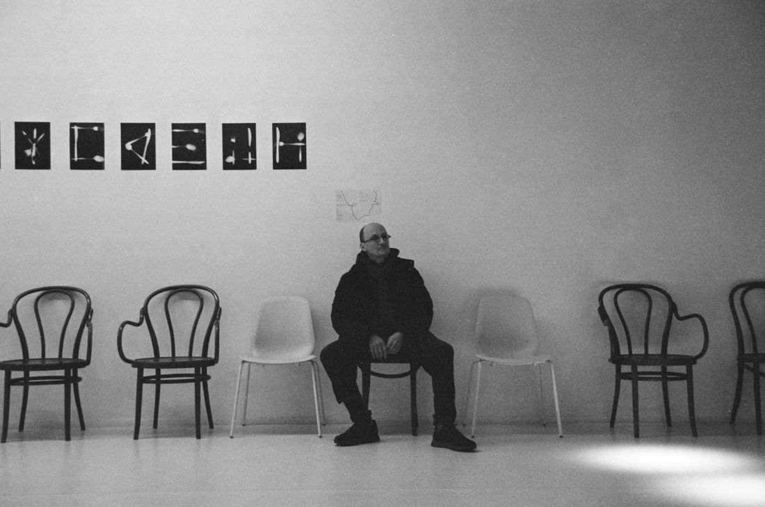 A single person seated among empty chairs in a plain room hung with small framed works