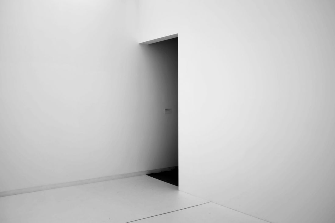 A wide, plain corner of an empty room with bare white walls and floor, a dark open doorway on one side.
