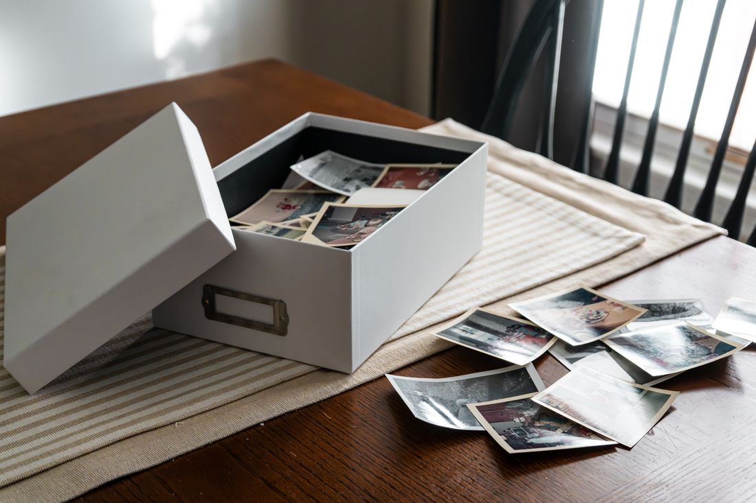 An open archive box of old photographs spread across a wooden table, the kind of material an anniversary film draws from