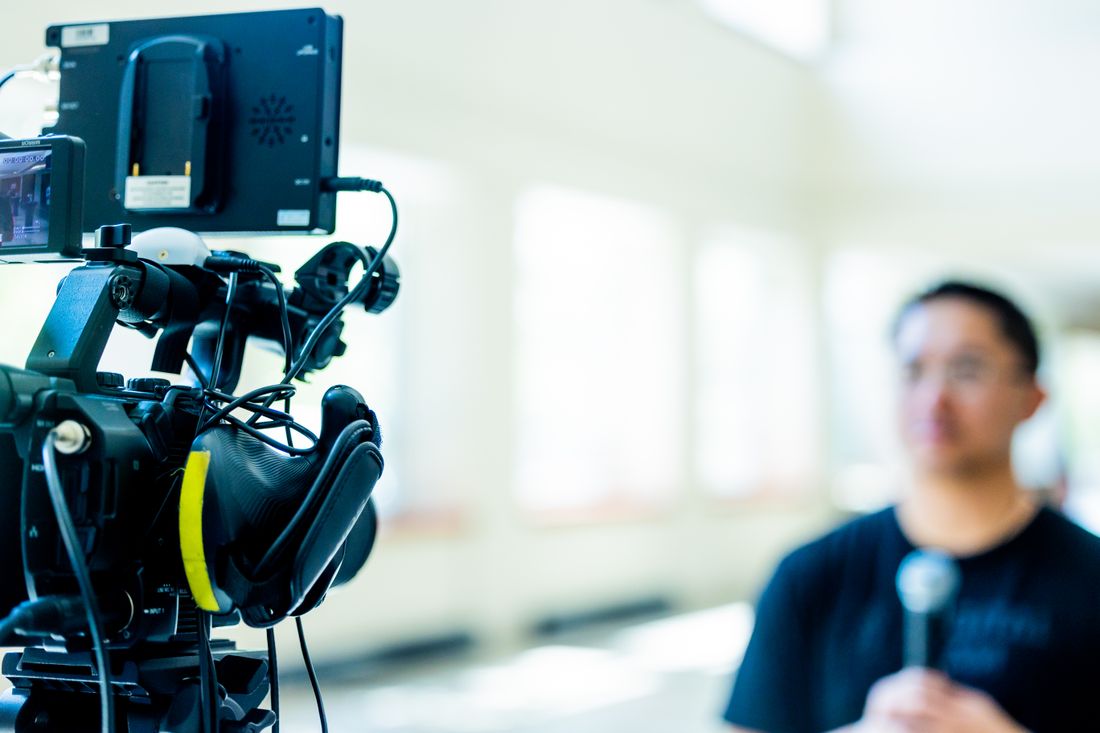 A video camera and monitor in focus on a tripod, with a blurred interview subject holding a microphone in the background