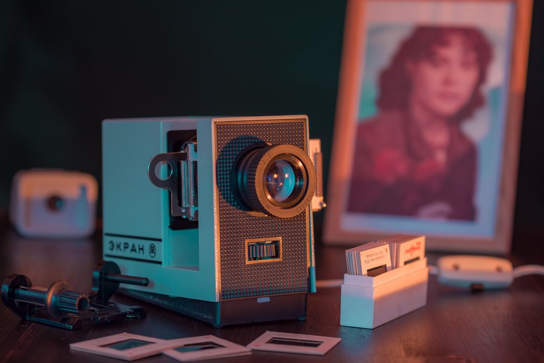 A vintage slide projector on a desk next to an old framed photograph, evoking the continuity between archive material and the present