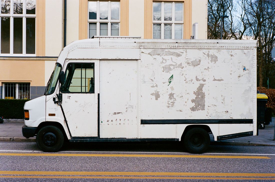 A worn white box truck parked at the curb in front of a plain building facade, no people visible.