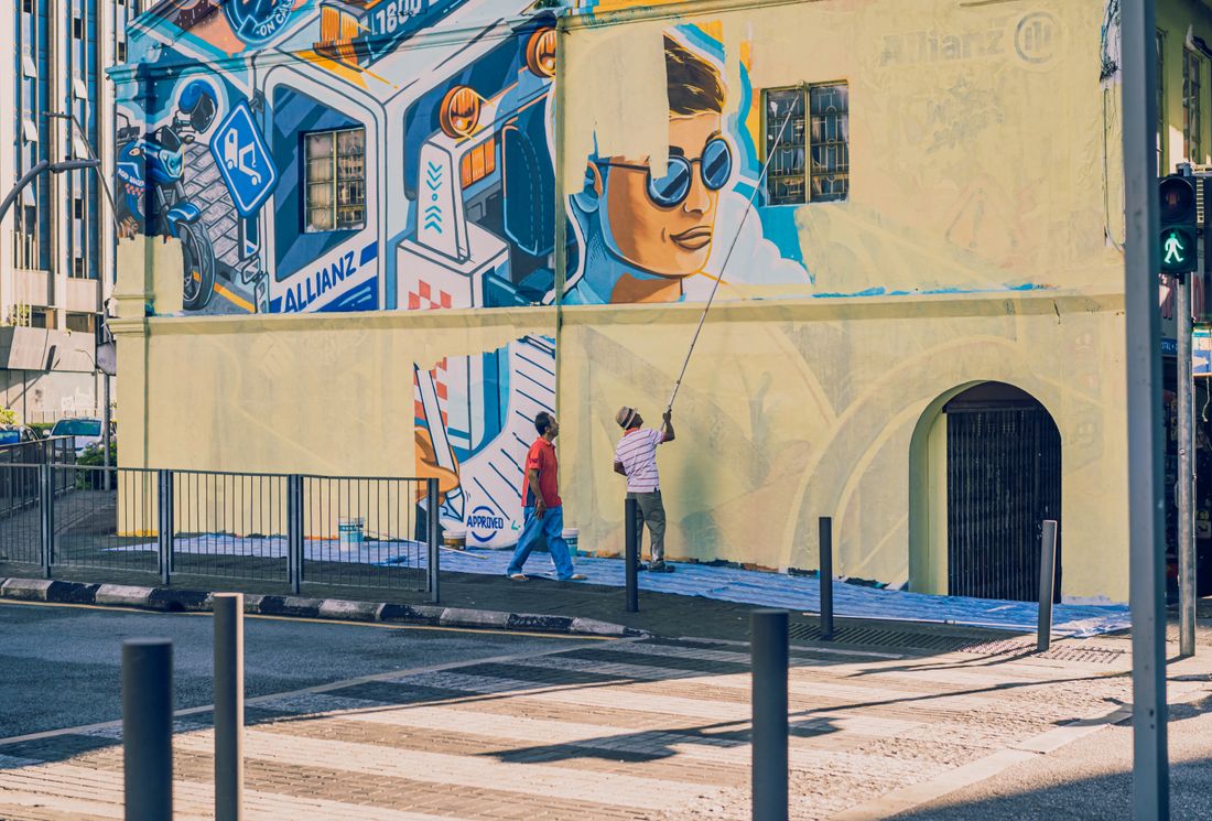 A person on a raised platform painting a large mural across the facade of a building, seen from across the street