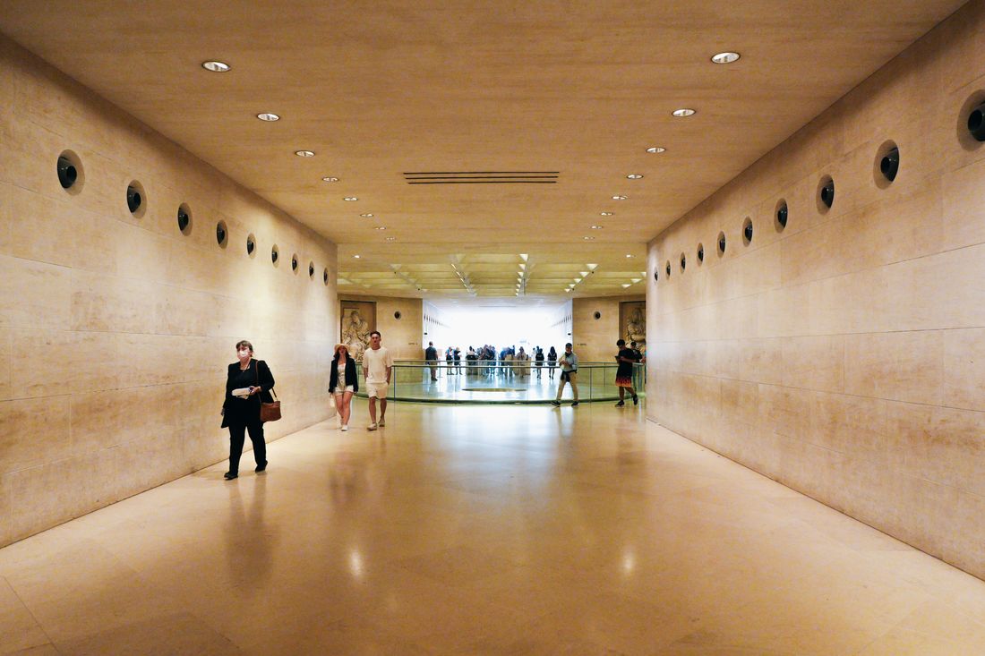 Museum visitors walking through a long stone corridor lined with wall niches and framed pieces.