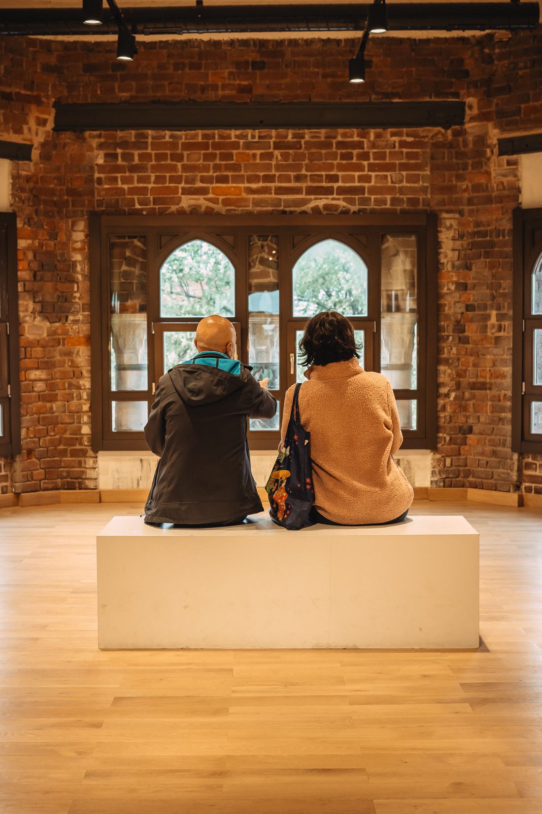 Two visitors seen from behind, seated together on a bench in a museum space with tall arched windows.