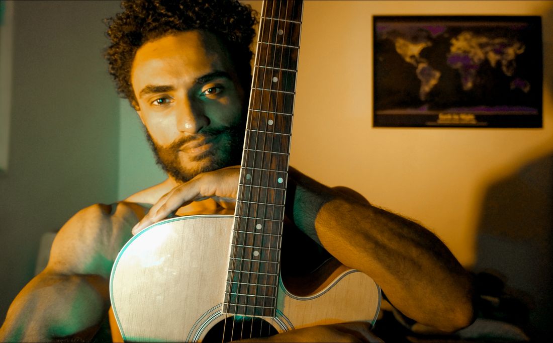 A musician sitting indoors, holding an acoustic guitar close in an intimate, warmly lit portrait