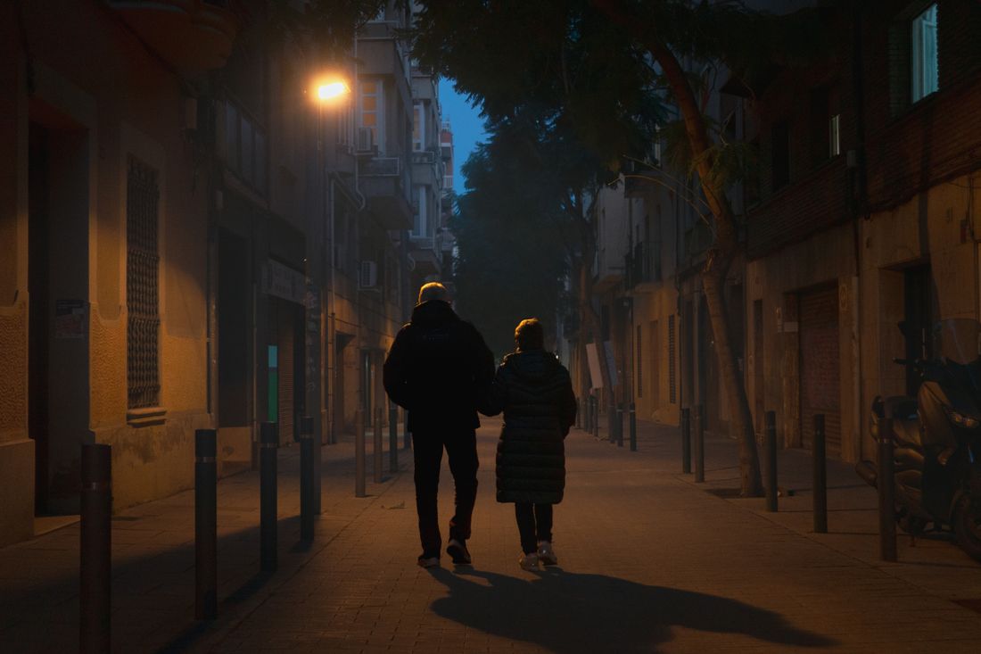 Silhouetted couple walking together on a quiet city street at night, lit by a single warm streetlamp against a deep blue evening sky.