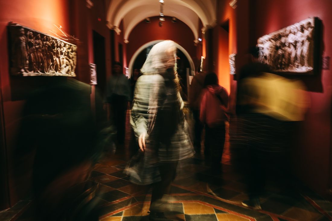 Motion-blurred crowd moving through a warmly lit arched gallery hallway, figures in soft focus beside framed wall pieces.