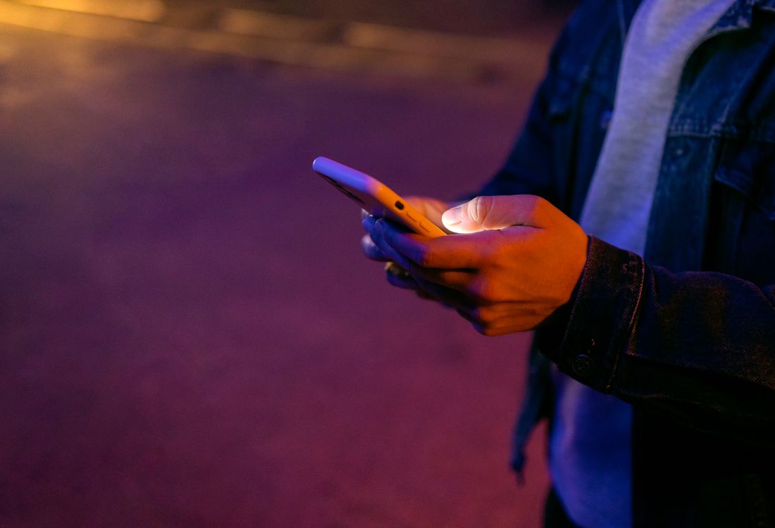 Close view of a person's hands holding a smartphone outdoors at night, lit by warm and violet ambient light, the screen not readable.