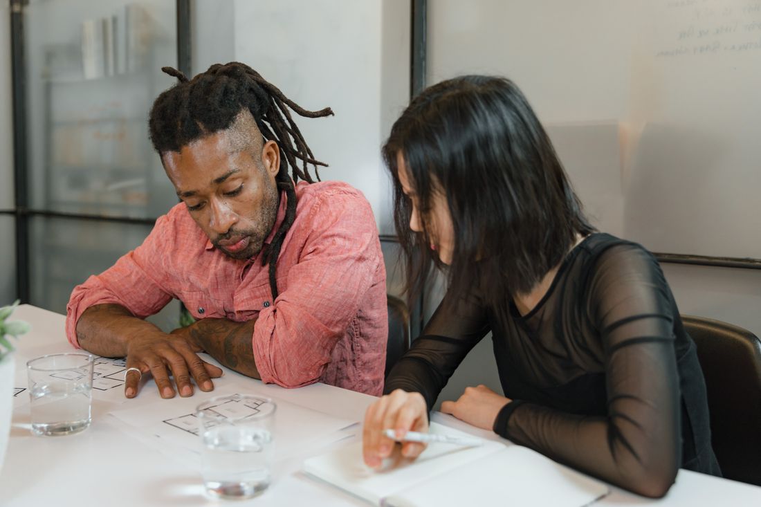 Two people seated at a table looking down together at a sketch on paper, one resting a hand near the page and the other writing, faces turned toward the page rather than the camera.
