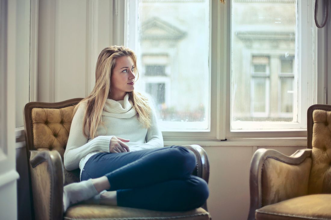 A woman seated in a cozy leather armchair by a window, hands folded, relaxed and unposed, plain wall and a second empty chair beside her.