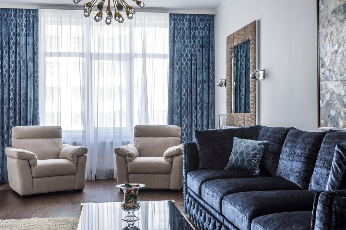An empty, softly furnished living room with velvet armchairs, heavy patterned curtains, sheer drapes, a rug, and natural daylight through the window.