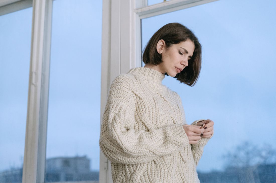 A woman standing near a window wearing a plain cream knit sweater, calm and unhurried, medium shot showing the outfit clearly.