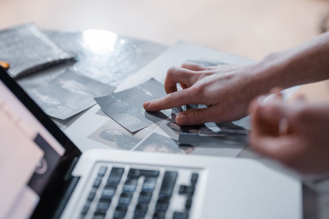 A hand sorting printed photographs on a table next to an open laptop, representing the file handoff after a shoot.