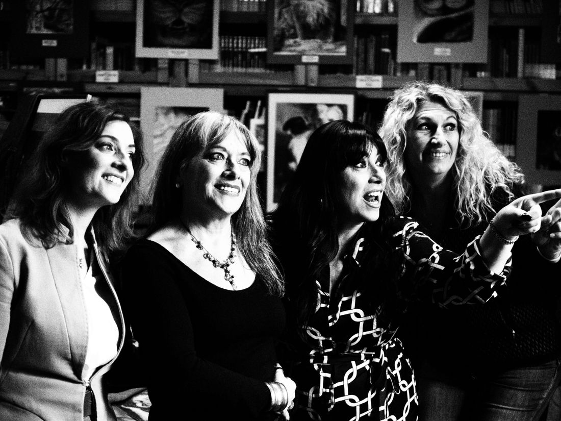 A small group of women in a gallery, standing close together and smiling near framed artwork on the wall.