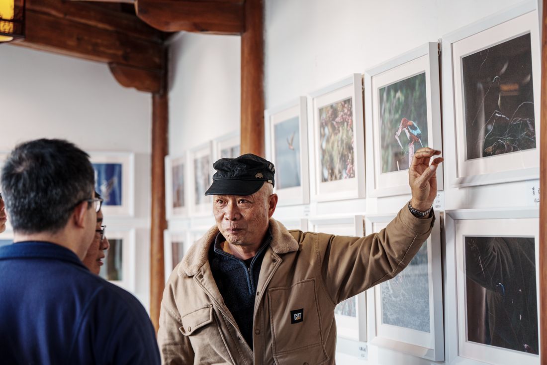 A gallery visitor gestures toward a row of framed photographs on the wall while two people listen nearby.