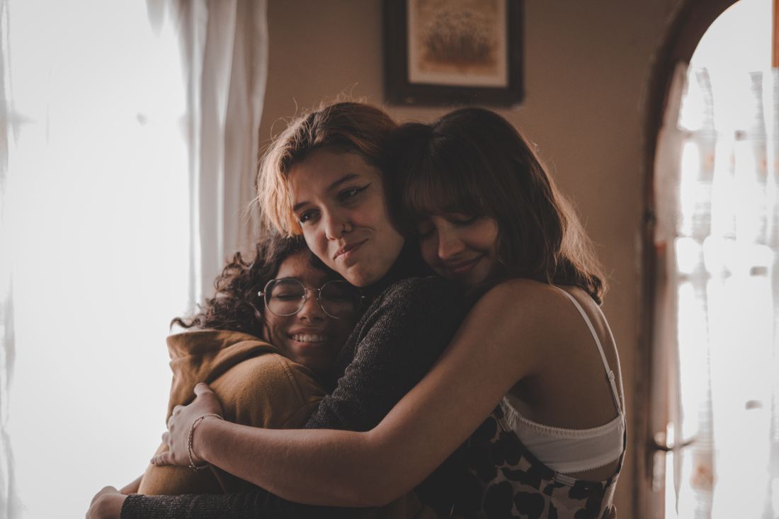 Three people share a warm group embrace indoors near a small framed picture on the wall, soft natural light.