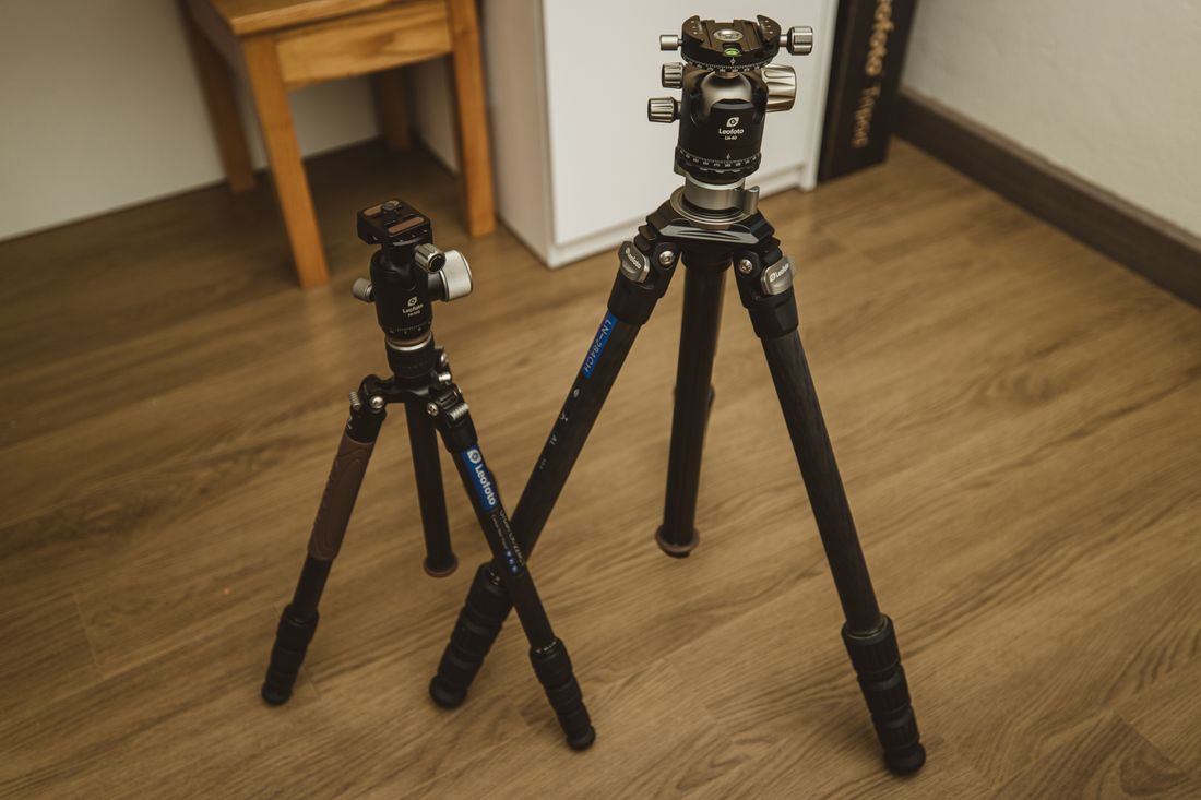 Two camera tripods of different sizes standing on a wooden floor