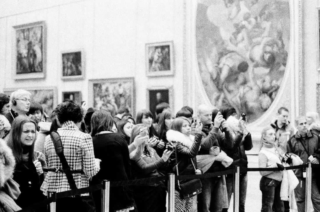 A crowd of people gathered closely to photograph an artwork in a gallery, seen from behind the group