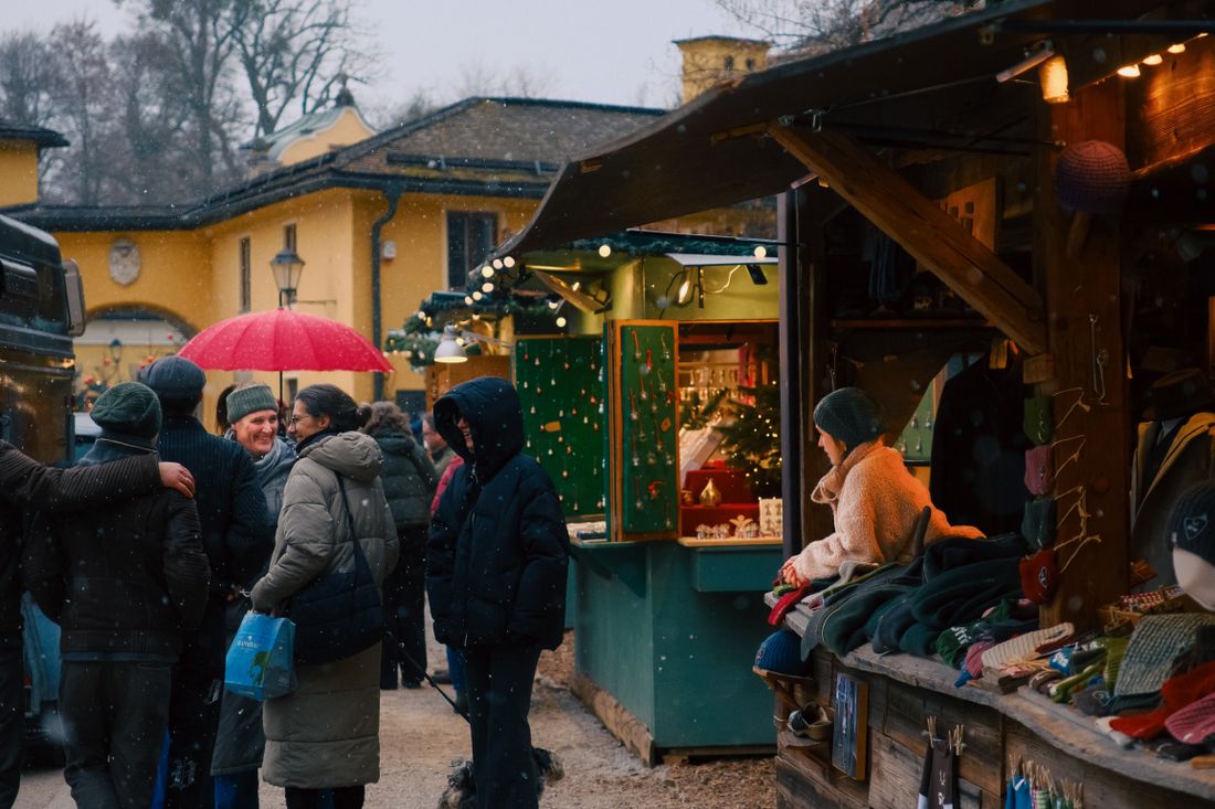 A warmly lit holiday market stall hung with ornaments, shoppers browsing nearby in the snow at dusk