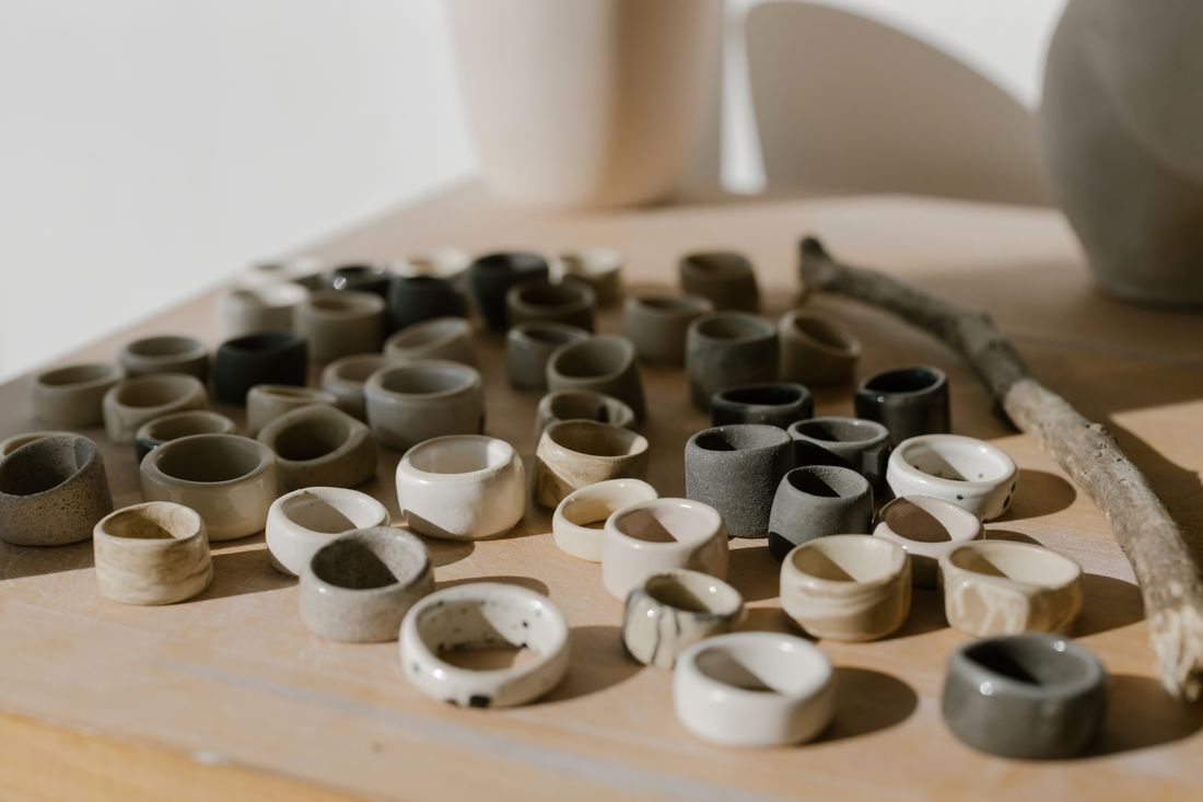 Dozens of handcrafted ceramic rings in mixed glaze colors arranged in neat rows on a wooden tabletop next to a bare branch