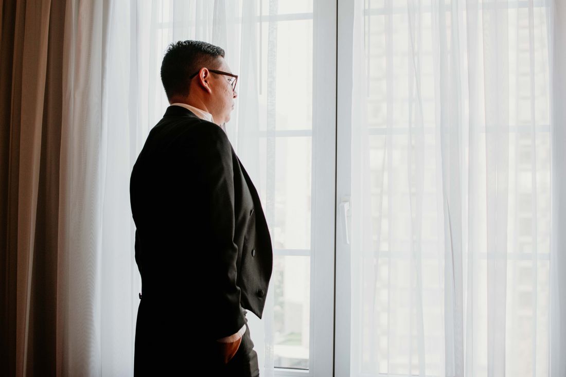 A man in a dark suit stands in profile beside a large window with sheer curtains, one hand in his pocket, looking out at a city view in soft daylight.